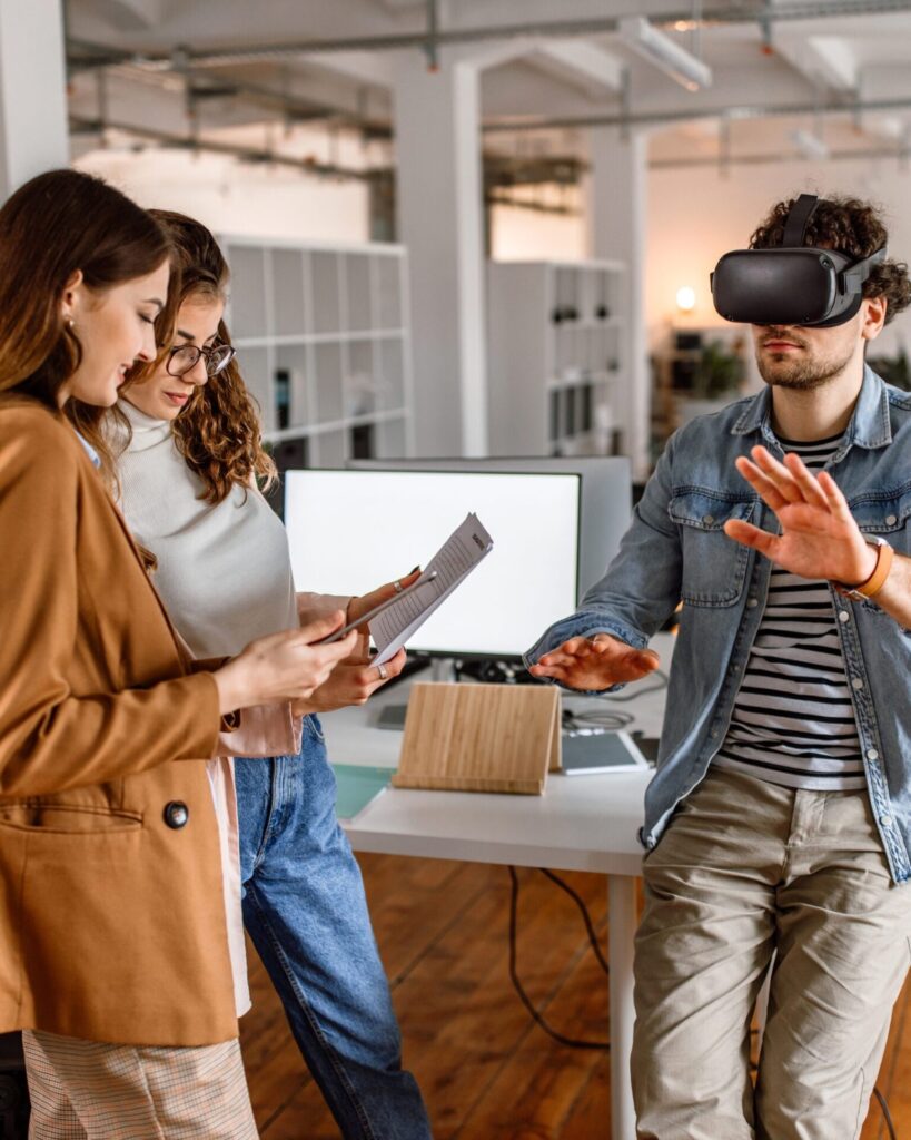 Cena: Escritório, homem com VR, mulheres com papel - Em um escritório moderno, um homem usa um headset de realidade virtual e gesticula, enquanto duas colegas ao lado analisam documentos, ilustrando a integração de RV em reuniões e treinamentos de equipe.